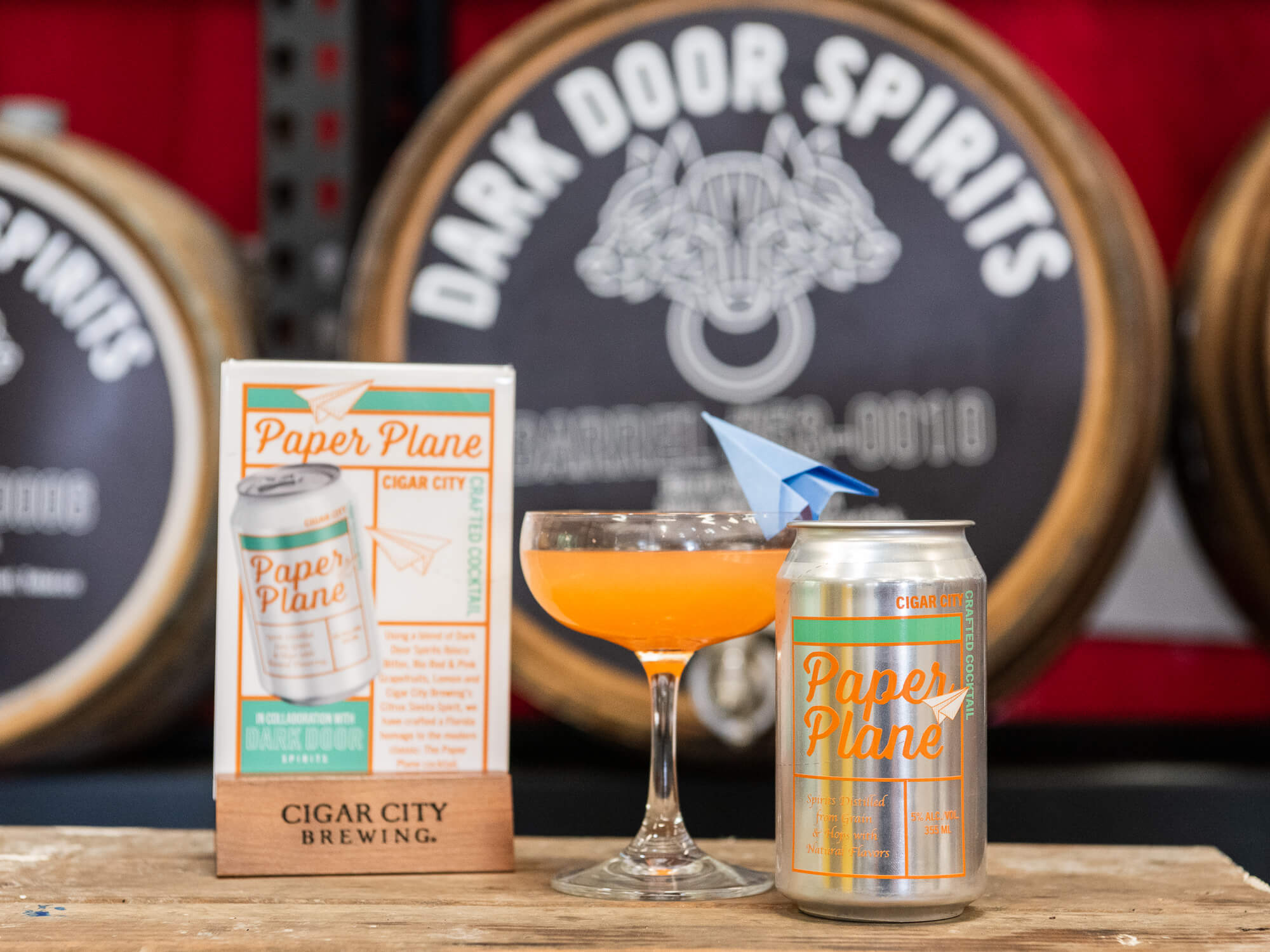 A glass of Paper Plane ready-to-drink cocktail sits next to its can in front of barrels baring the Dark Door Spirits brand imagery.