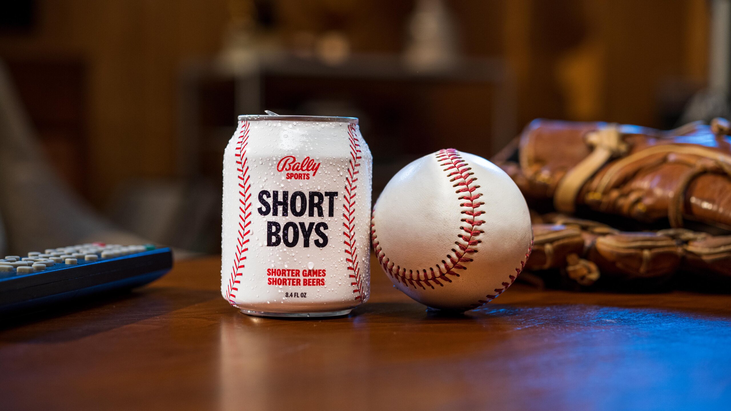 a shorter than average can of beer sits on a table next to a baseball, with a TV remote control and a baseball glove in the background. The can says Bally Sports Short Boys; Shorter Games, Shorter Beers 8.4 FL OZ